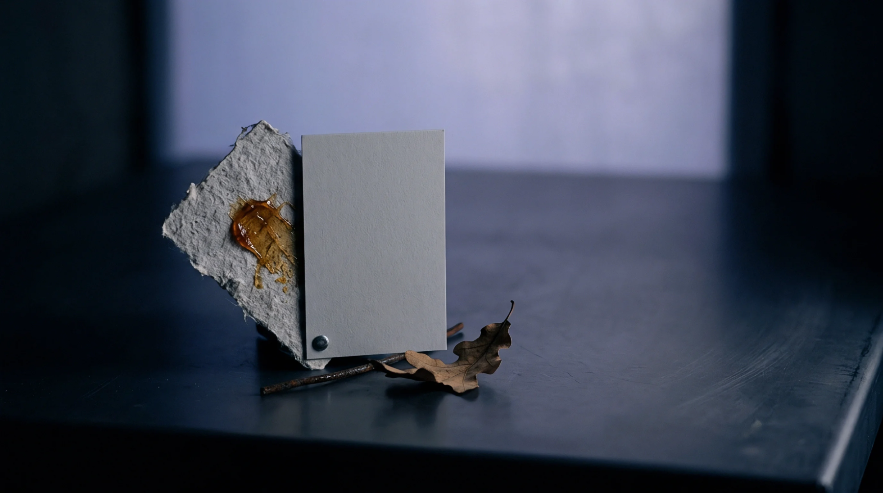 A smooth white card secured by a metal fastener to a textured recycled substrate features a glossy amber coating beside an isolated dried oak leaf.
