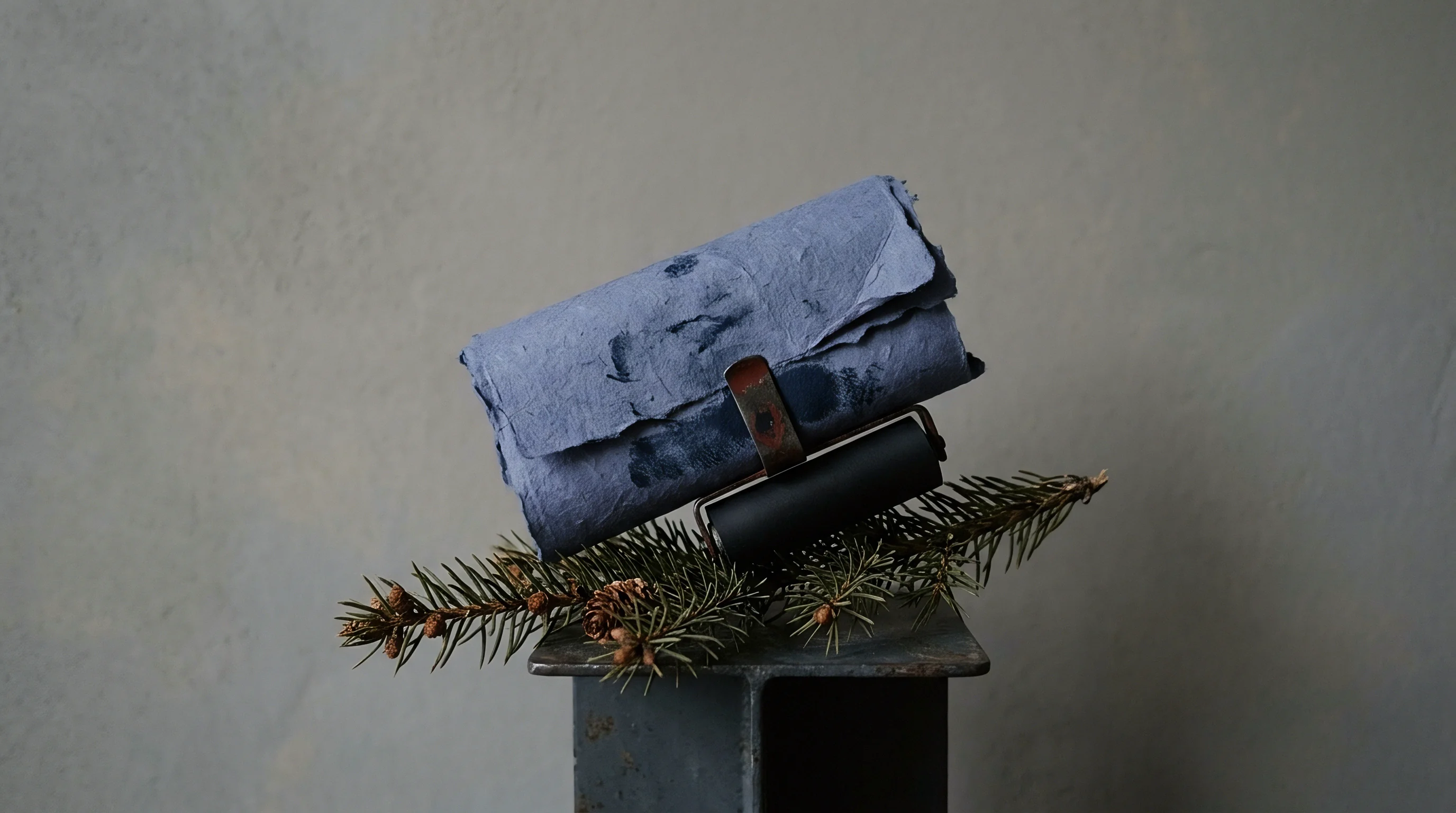 A heavy blue fibrous paper sheet rests in a metal brayer frame above pine needles on a dark industrial steel pedestal.