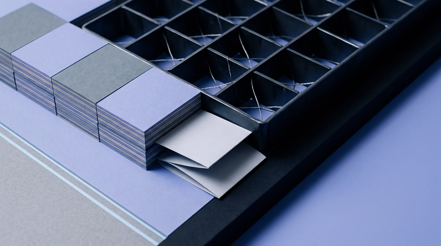 Neatly stacked squares of lavender and grey cardstock are positioned beside a compartmentalized grid tool containing loose paper elements.