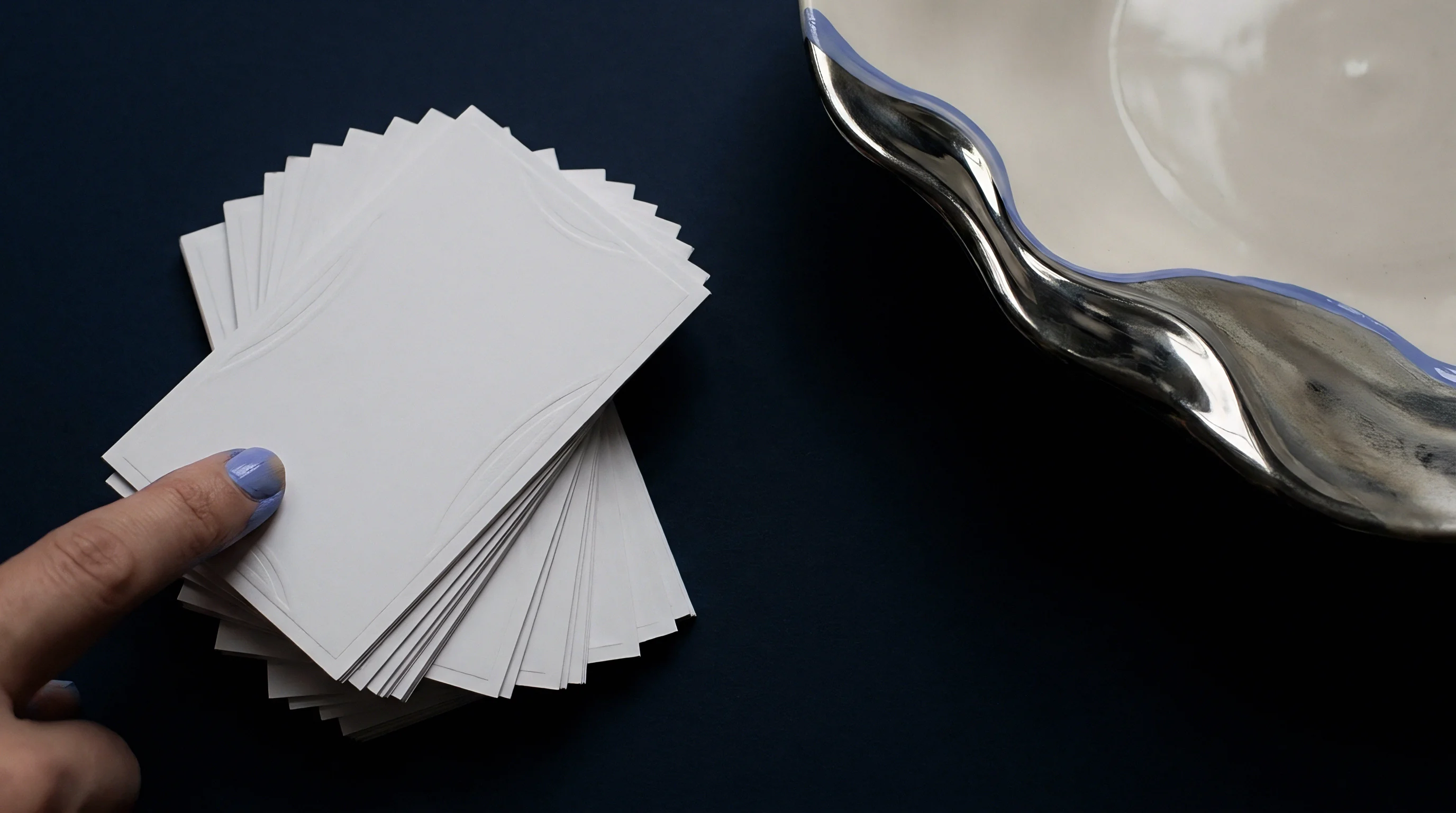 A finger with blue polish touches a fanned stack of blank white cardstock substrates resting on a dark navy surface.